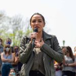 Woman speaking into a microphone outdoors, crowd behind.