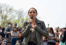 Woman speaking into a microphone outdoors, crowd behind.