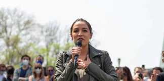 Woman speaking into a microphone outdoors, crowd behind.