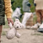 Child holding teddy bear near people and tent outside.