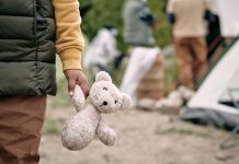 Child holding teddy bear near people and tent outside.