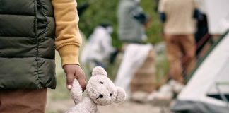 Child holding teddy bear near people and tent outside.