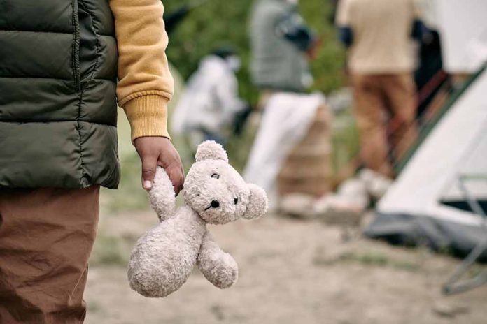 Child holding teddy bear near people and tent outside.