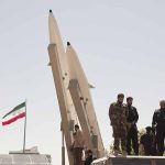 Military personnel beside missiles and Iranian flag.