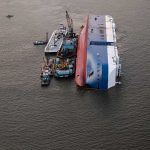 Capsized ship with cranes and tugboats around it.