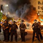 Police officers in riot gear near burning car.