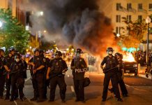 Police officers in riot gear near burning car.