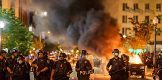 Police officers in riot gear near burning car.