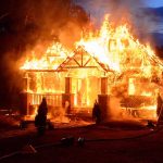 House engulfed in flames with firefighters present.