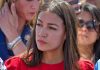 Woman in red shirt with serious expression, background blurry.