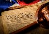 Gavel on U.S. Constitution with flag backdrop.