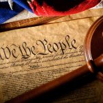 Gavel on U.S. Constitution with flag backdrop.