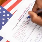 Hand filling out voter registration form next to flag.