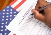 Hand filling out voter registration form next to flag.