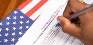 Hand filling out voter registration form next to flag.