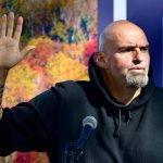 Man in black hoodie raising hand, speaking at microphone.