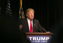 Man speaking at podium with TRUMP sign