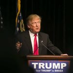 Man speaking at podium with TRUMP sign