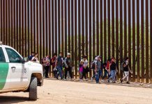 People walking beside tall fence and border patrol vehicle