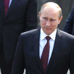 Putin Reveals Parades Nuke – Global Panic! A serious-looking man in a suit walking outdoors with security personnel in the background