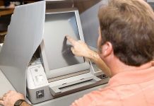 Election Computer FLIPS Mid-term Winner After Taking Office Person using touchscreen voting machine in polling booth.
