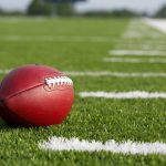 Legend Coach RUSHED to Hospital – Shot at College! A red American football resting on a green artificial turf field