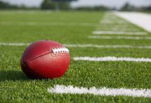 Legend Coach RUSHED to Hospital – Shot at College! A red American football resting on a green artificial turf field