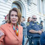 Shocking Power Void Opens – Pelosi’s Seat Up For Grabs A woman in an orange blazer speaking passionately at a rally with security personnel in the background
