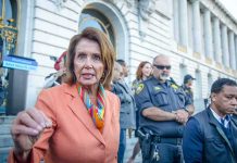 Shocking Power Void Opens – Pelosi’s Seat Up For Grabs A woman in an orange blazer speaking passionately at a rally with security personnel in the background