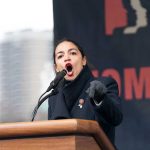Woman speaking passionately at podium during outdoor event.