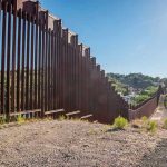 Tall metal border wall with rural landscape.