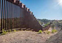 U.S Sets Up MILITARIZED Zone Along Border Tall metal border wall with rural landscape.