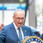 Schumer’s Shutdown Gamble – America Exposed! Man in suit and glasses speaks passionately at podium.
