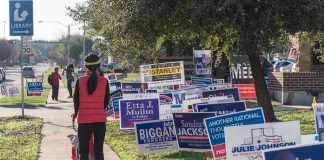 Senate SHOCKWAVE STUNS Republican Elite Person walking with stroller past numerous election campaign signs.