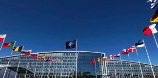 Real Reason Behind US Military Moves in Europe Flags outside NATO headquarters building under clear blue sky.
