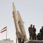 Report of Resignation Triggers Questions Over Who Runs the State Military personnel beside missiles and Iranian flag.