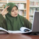 Student in green hijab studying with laptop and books in a library