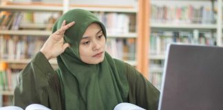 Student in green hijab studying with laptop and books in a library