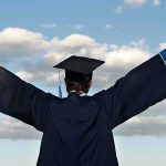 Graduate in cap and gown with arms raised against a blue sky