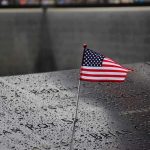 9/11 Families and NYC Mayor Clash Ahead of 25th Anniversary American flag on a rainy memorial nameplate.