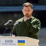 A public figure in a green sweater pondering during a speech at a podium in Kyiv
