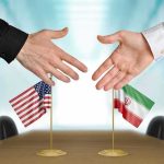 Two hands reaching to shake above US and Iran flags on a conference table