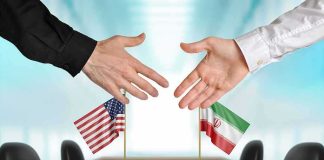 Two hands reaching to shake above US and Iran flags on a conference table