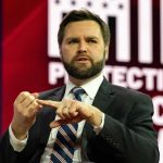 Governor Race Turns Ugly: What’s the Real Story? Man in suit speaking on stage with red and white backdrop