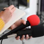 California’s New Tax Squad Just Picked a Celebrity Journalist holding two microphones while writing in a notebook
