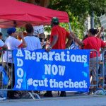 Reparations Proposals Face Legal Questions Protesters behind barricades holding a blue reparations banner