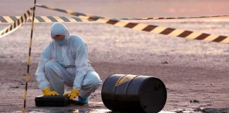 Hazmat worker kneeling near a fallen barrel at a cordoned-off site