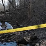 Grapple Game Ends In Manslaughter forensic investigators in white suits examine a forested stream within taped-off area