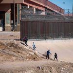 People walking along a dry canal beside a high fence with barbed wire