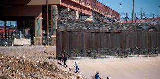 People walking along a dry canal beside a high fence with barbed wire
